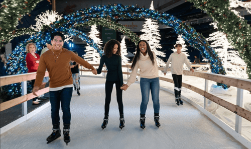A group of friends ice skating together, smiling and holding hands, surrounded by festive lights and decorations.