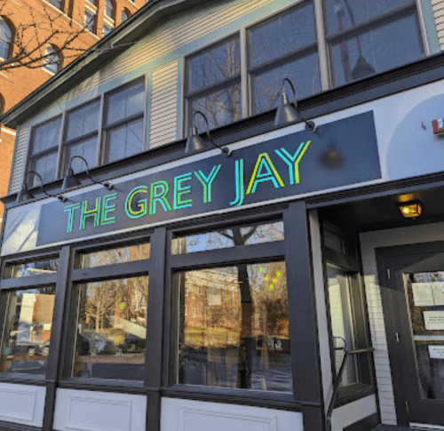 Sign for "The Grey Jay" restaurant, featuring bright green lettering on a building with large windows.
