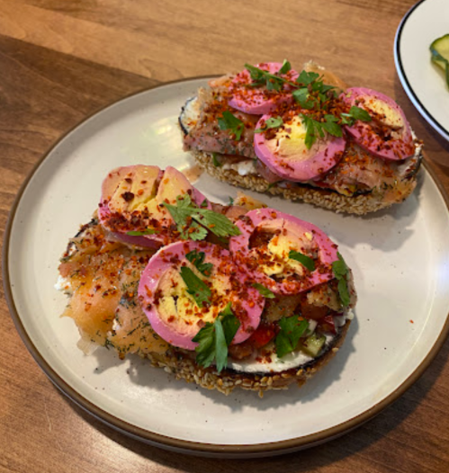 Two open-faced sandwiches topped with smoked salmon, pickled onions, herbs, and spices on a wooden table.