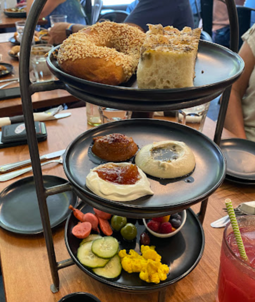 A tiered serving stand with bagels, focaccia, dips, vegetables, and fruits on black plates, set on a wooden table.