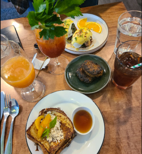 A table set with brunch items: French toast, eggs Benedict, drinks, and a small dessert on a wooden surface.