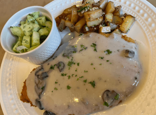 A plate with chicken fried steak topped with mushroom gravy, served with diced potatoes and a cucumber salad.