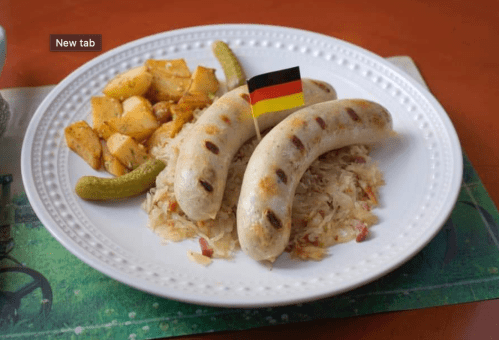 Plate of grilled sausages with a small German flag, served with sauerkraut, roasted potatoes, and pickles.