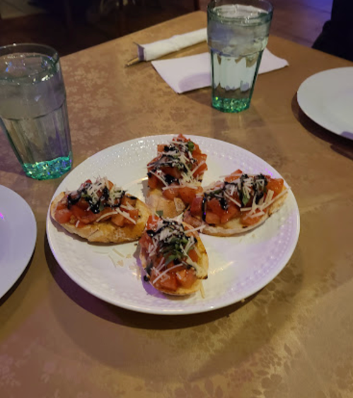 A plate of bruschetta topped with diced tomatoes, cheese, and balsamic drizzle, served with glasses of water.