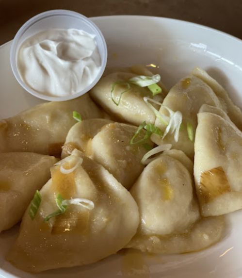 A bowl of dumplings drizzled with sauce, garnished with green onions, served with a side of sour cream.