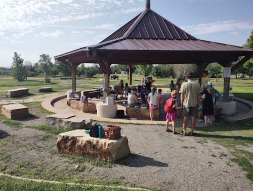 A group of people gathers under a gazebo in a park, with picnic tables and grassy areas around them.