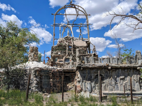 A unique, rustic structure made of stone and wood, surrounded by a fence and set against a blue sky with clouds.