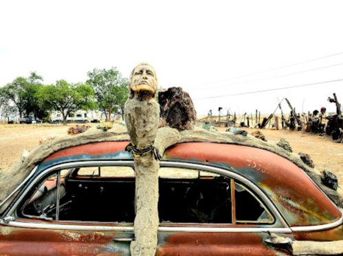 A weathered car with a sculpted figure on top, set in a barren landscape with sparse trees in the background.