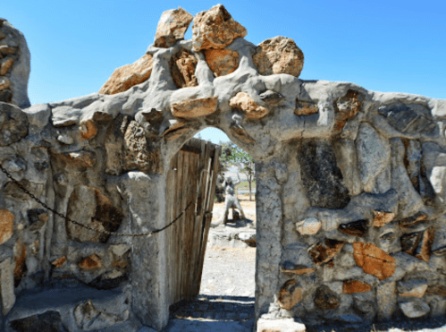 A weathered stone archway with a wooden gate, leading to a rocky landscape under a clear blue sky.