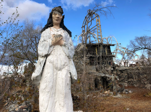 A weathered statue of a woman in white stands in front of a dilapidated building surrounded by bare trees.
