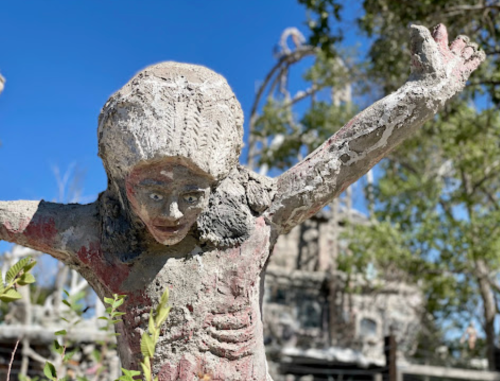 A weathered statue of a child with outstretched arms, surrounded by greenery and a clear blue sky.