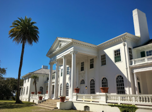 A grand white mansion with columns, surrounded by palm trees and a clear blue sky.