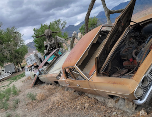A cluster of vintage cars, partially buried and rusted, with a statue in the background against a cloudy sky.