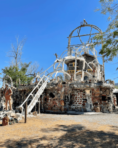 A whimsical, multi-textured building with a spiral staircase, surrounded by sculptures and greenery under a clear blue sky.