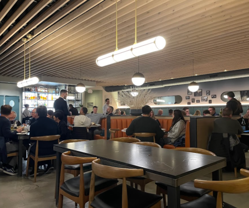 A modern restaurant interior with diners at tables, warm lighting, and a bar in the background.