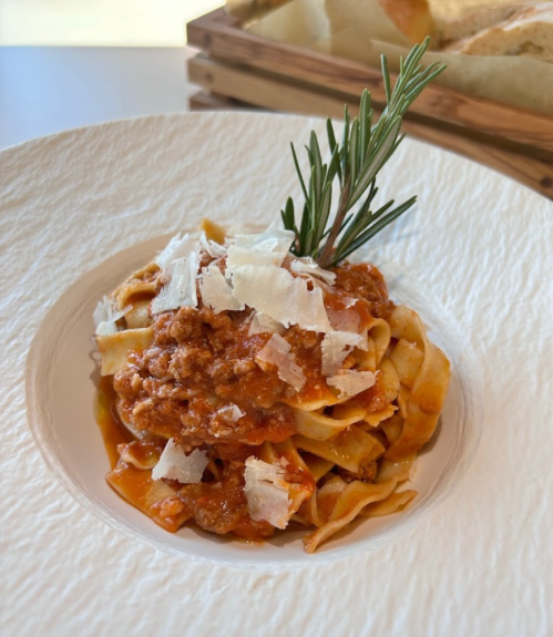 A plate of pasta with meat sauce, topped with shaved cheese and a sprig of rosemary, served on a textured white dish.