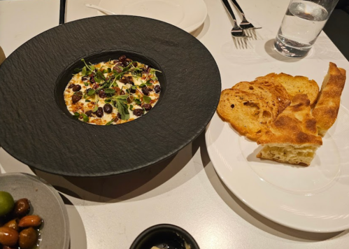 A black plate with a creamy dish topped with herbs and nuts, accompanied by toasted bread and olives on the side.