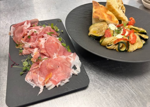 A platter of sliced cured meats with herbs, alongside a salad of grilled vegetables and bread on a dark plate.
