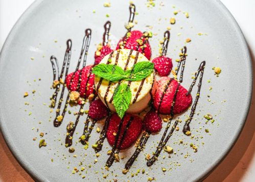 A dessert featuring a round cake topped with mint, surrounded by raspberries and drizzled with chocolate and pistachios.