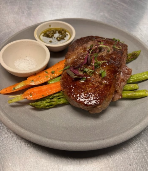 A plated steak with asparagus and carrots, accompanied by small bowls of sauce and salt.