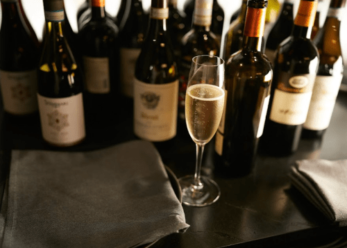A glass of sparkling wine on a table with various wine bottles in the background. Two gray napkins are nearby.