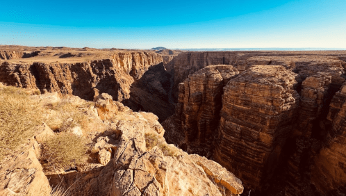 A vast canyon landscape with steep rock formations and a clear blue sky above.