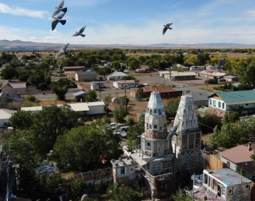 Aerial view of a quirky, castle-like structure surrounded by trees and houses, with birds flying overhead.