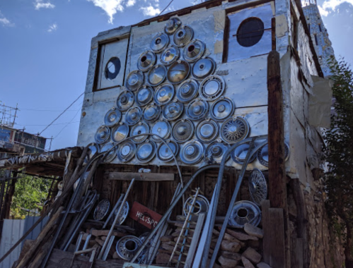 A quirky building adorned with hubcaps arranged in a tree shape, surrounded by rustic materials and blue sky.
