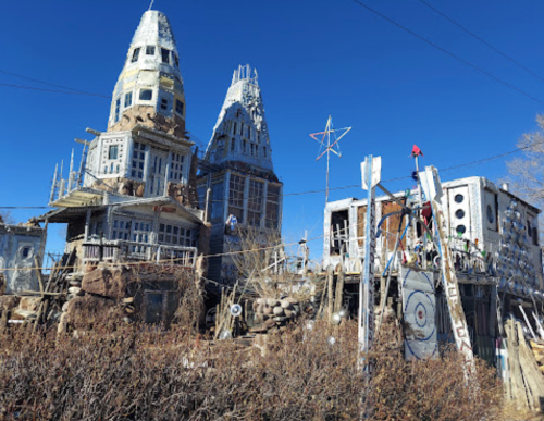 Whimsical, multi-towered structure with eclectic design, surrounded by rocky terrain and clear blue sky.