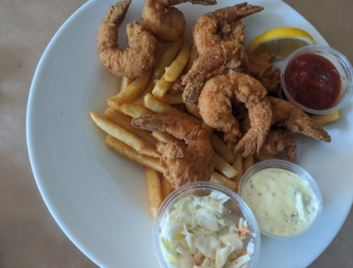 A plate of fried shrimp with French fries, coleslaw, and dipping sauces, garnished with a lemon wedge.