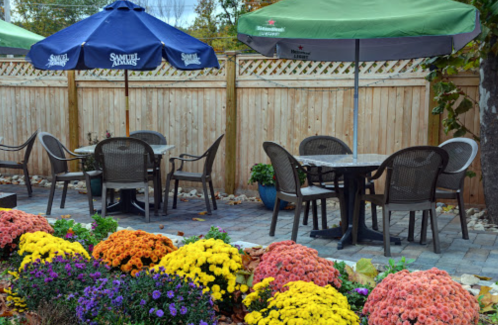 Outdoor seating area with tables under umbrellas, surrounded by colorful flowers and a wooden fence.
