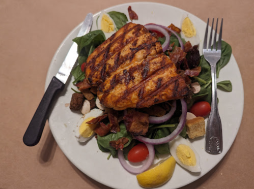 Grilled salmon on a bed of spinach, topped with bacon, tomatoes, onions, croutons, and hard-boiled eggs, with lemon.
