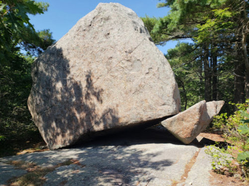 A large boulder balanced on a smaller rock, surrounded by trees and greenery in a natural setting.