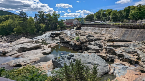 A scenic view of a rocky river with a dam, surrounded by trees and a blue sky with fluffy clouds.
