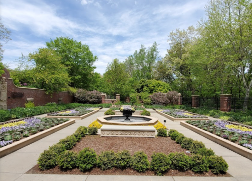 A landscaped garden featuring a central fountain, colorful flower beds, and lush greenery under a blue sky.