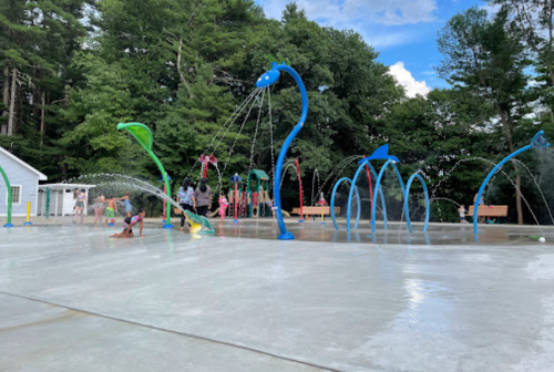 Children play in a colorful splash pad with water features, surrounded by trees and a playground in the background.