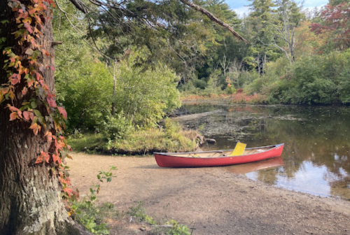 A red canoe rests on a sandy shore beside a calm, reflective pond surrounded by lush greenery and autumn foliage.