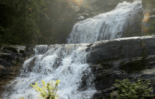 A cascading waterfall flows over rocky terrain, surrounded by lush greenery and dappled sunlight.