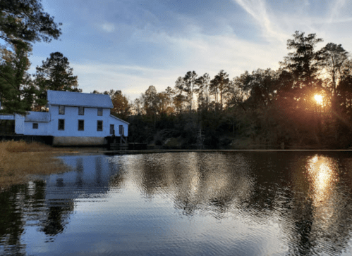 A serene sunset over a calm lake, reflecting a white house surrounded by trees.