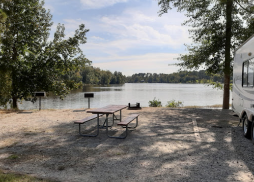 A serene lakeside campsite with a picnic table, grills, and an RV, surrounded by trees and calm water.