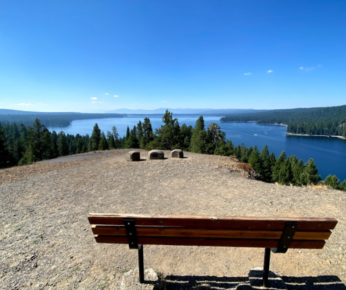 A wooden bench overlooks a serene lake surrounded by trees and mountains under a clear blue sky.