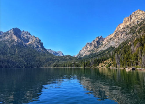 A serene lake surrounded by mountains and lush forests under a clear blue sky. Reflections shimmer on the water's surface.