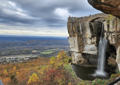 A waterfall cascades from a rocky cliff, surrounded by autumn foliage and a scenic valley under a cloudy sky.