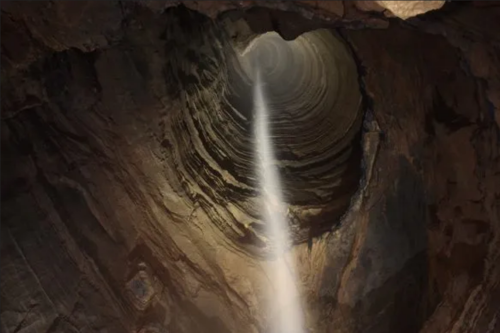 A deep, cylindrical cave with a beam of light shining down from the top, illuminating the rocky walls.
