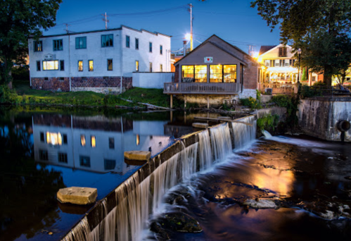 A serene evening scene of a small waterfall beside buildings, reflecting lights on calm water.