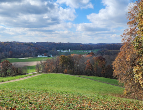 A scenic view of rolling green hills, autumn trees, and a distant farm under a partly cloudy sky.