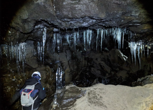 A person in a helmet explores a dark cave with icicles hanging from the rocky ceiling.