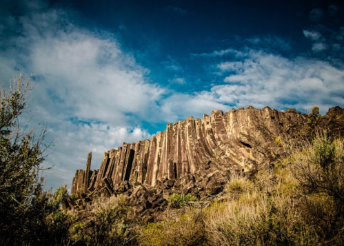 A rocky cliff with tall, columnar formations under a blue sky with scattered clouds and sparse vegetation in the foreground.