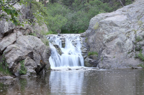A serene waterfall cascades over rocks into a calm pool, surrounded by lush greenery and trees.