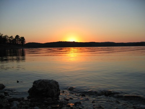 Sunset over a calm lake, with vibrant orange and yellow hues reflecting on the water's surface and rocky shoreline.